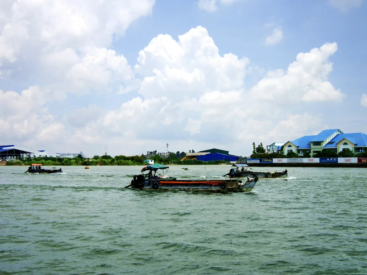 Những chiếc thuyền gỗ nhỏ băng qua sông rộng ở Đồng Tháp, phía xa là nhà máy mái xanh ven bờ.