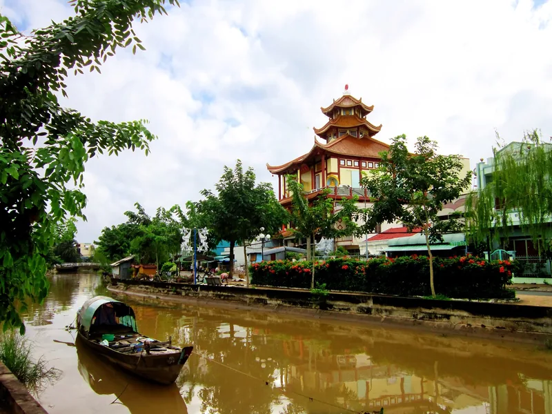 Một chiếc thuyền gỗ trôi trên con rạch ở Sa Đéc, bên cạnh ngôi chùa nhiều màu sắc và cây xanh tươi tốt.