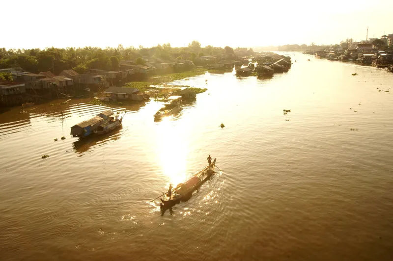 Một chiếc xuồng nhỏ lướt qua sông Mekong vàng óng gần những căn nhà sàn ven sông lúc hoàng hôn ở Châu Đốc.