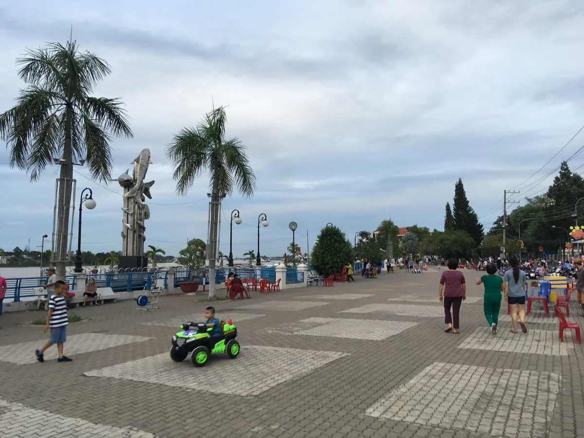 Con đường ven sông ở Châu Đốc với hàng cây cọ, tượng cá và người dân đi dạo buổi chiều.
