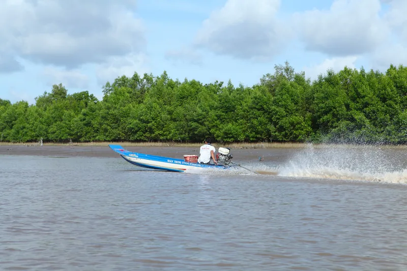 Một chiếc xuồng máy lướt nhanh tạo bọt nước trên sông, phía sau là rừng ngập mặn gần Cà Mau.